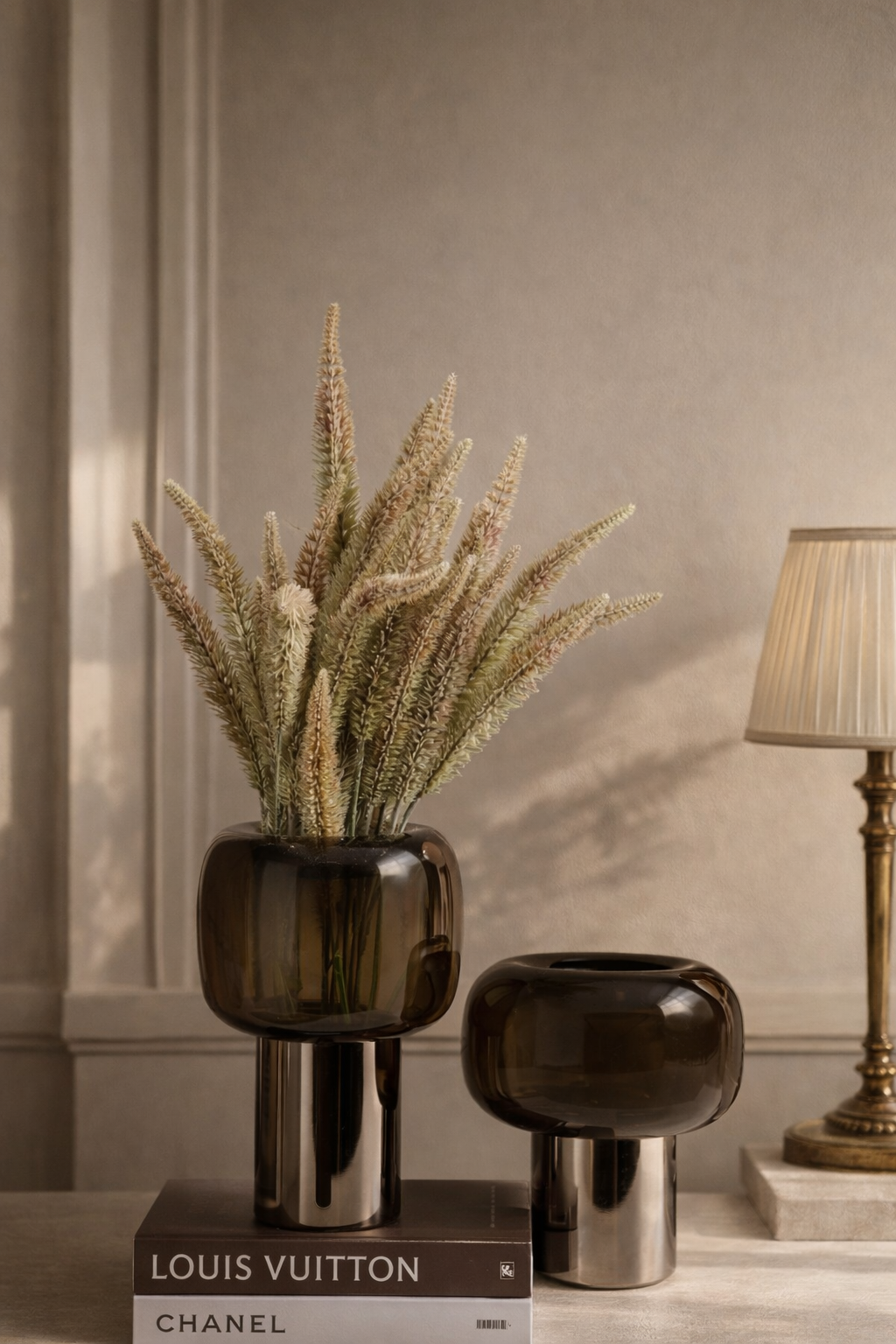 Decorative vases with a plant on a book titled 'Louis Vuitton' against a beige wall.