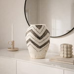Decorative vase with chevron pattern on a white surface, with a mirror and candle in the background.