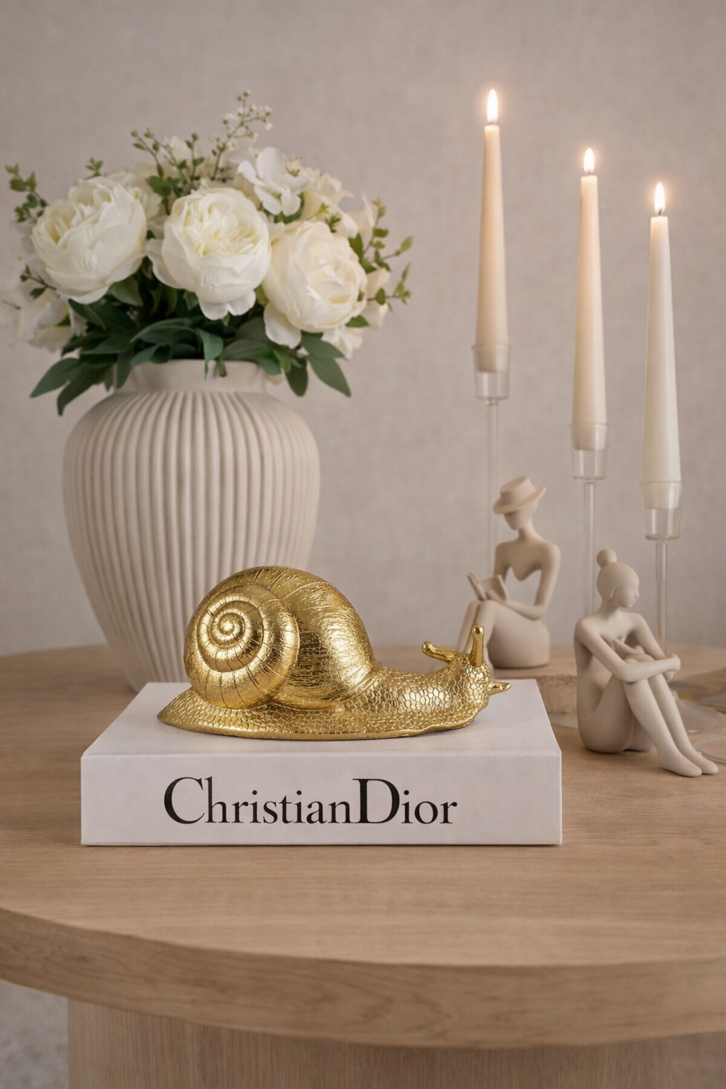 Decorative setup with a gold snail sculpture on a 'Christian Dior' book, vase with flowers, and candles.