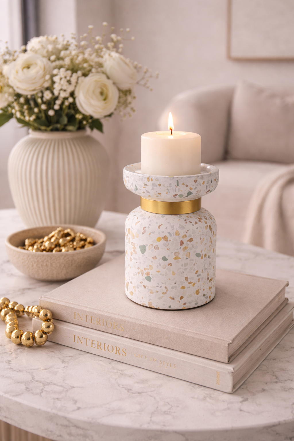 Decorative candle holder with a candle on top of books, surrounded by flowers and a bowl on a marble surface.