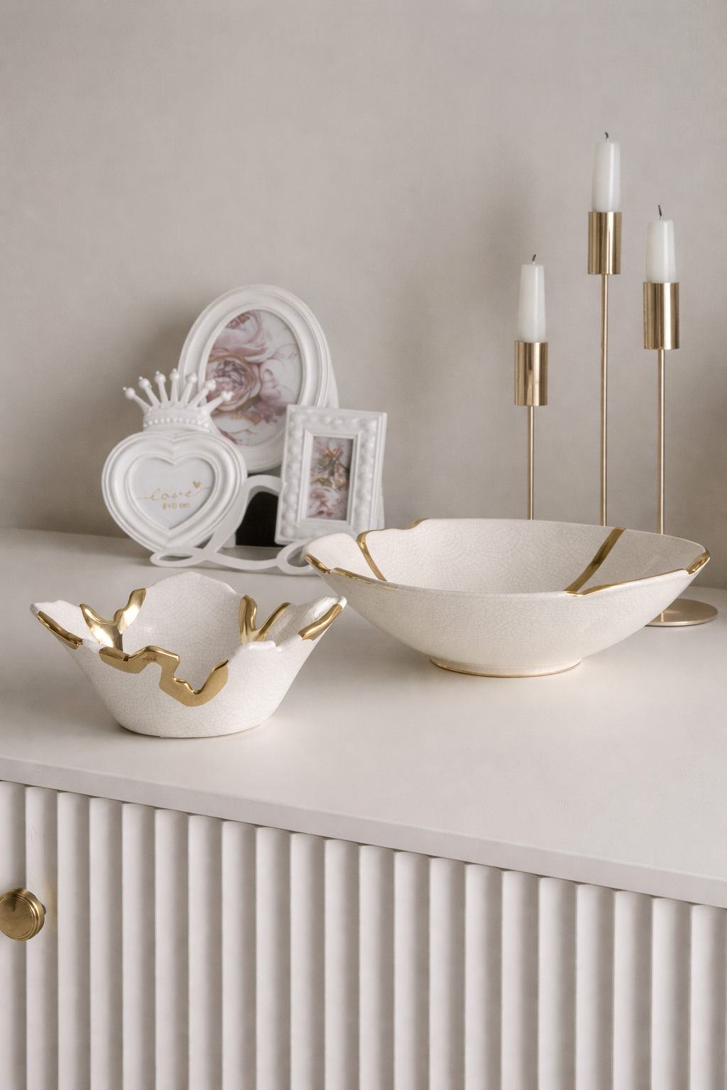 Decorative white bowls with gold accents on a shelf with candles and photo frames.