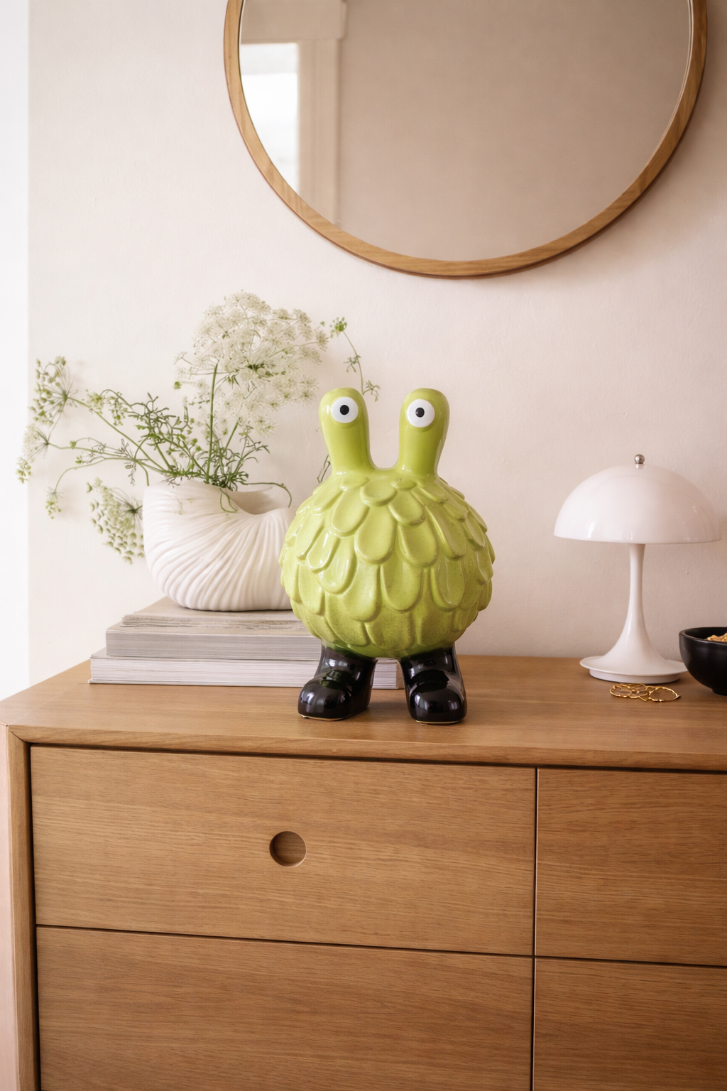 Green ceramic artichoke-shaped object on a wooden dresser with a mirror and lamp in the background.