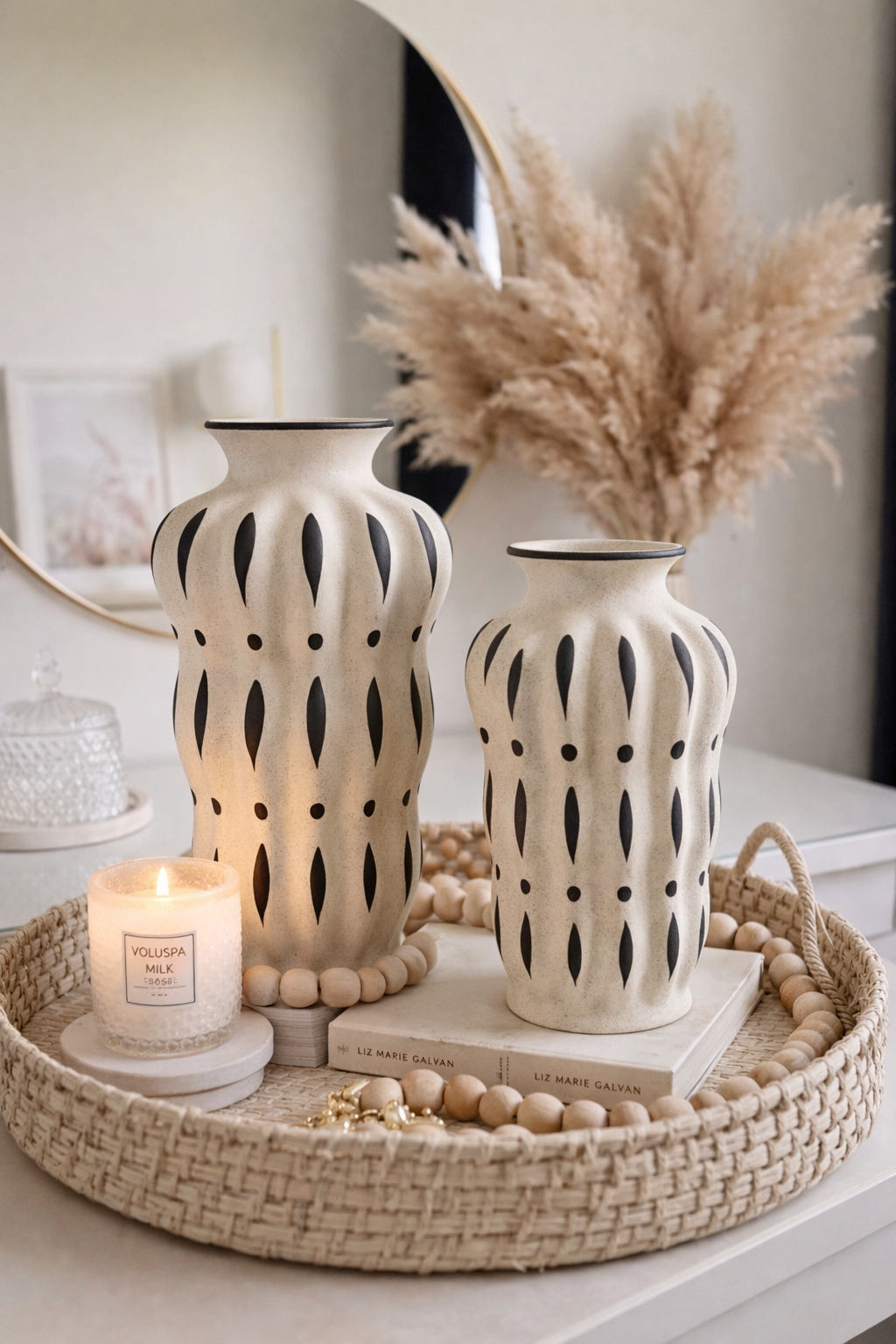 Decorative setting with two patterned vases, a candle, and dried pampas grass on a woven tray.