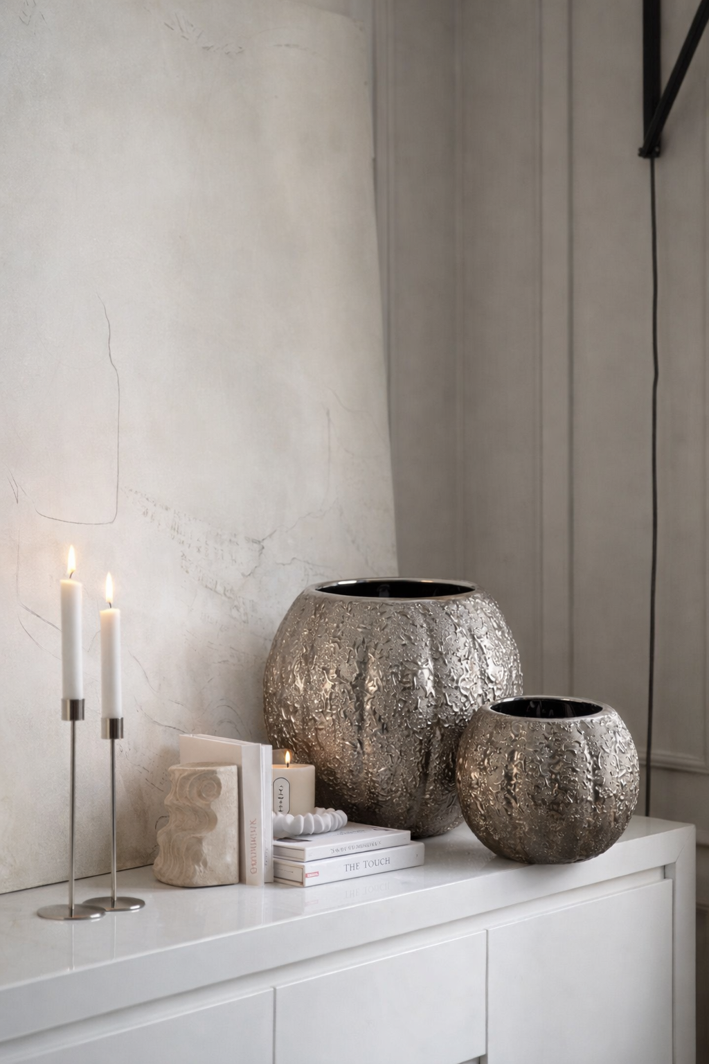 Decorative silver vases on a white surface with candles and books against a light gray wall.