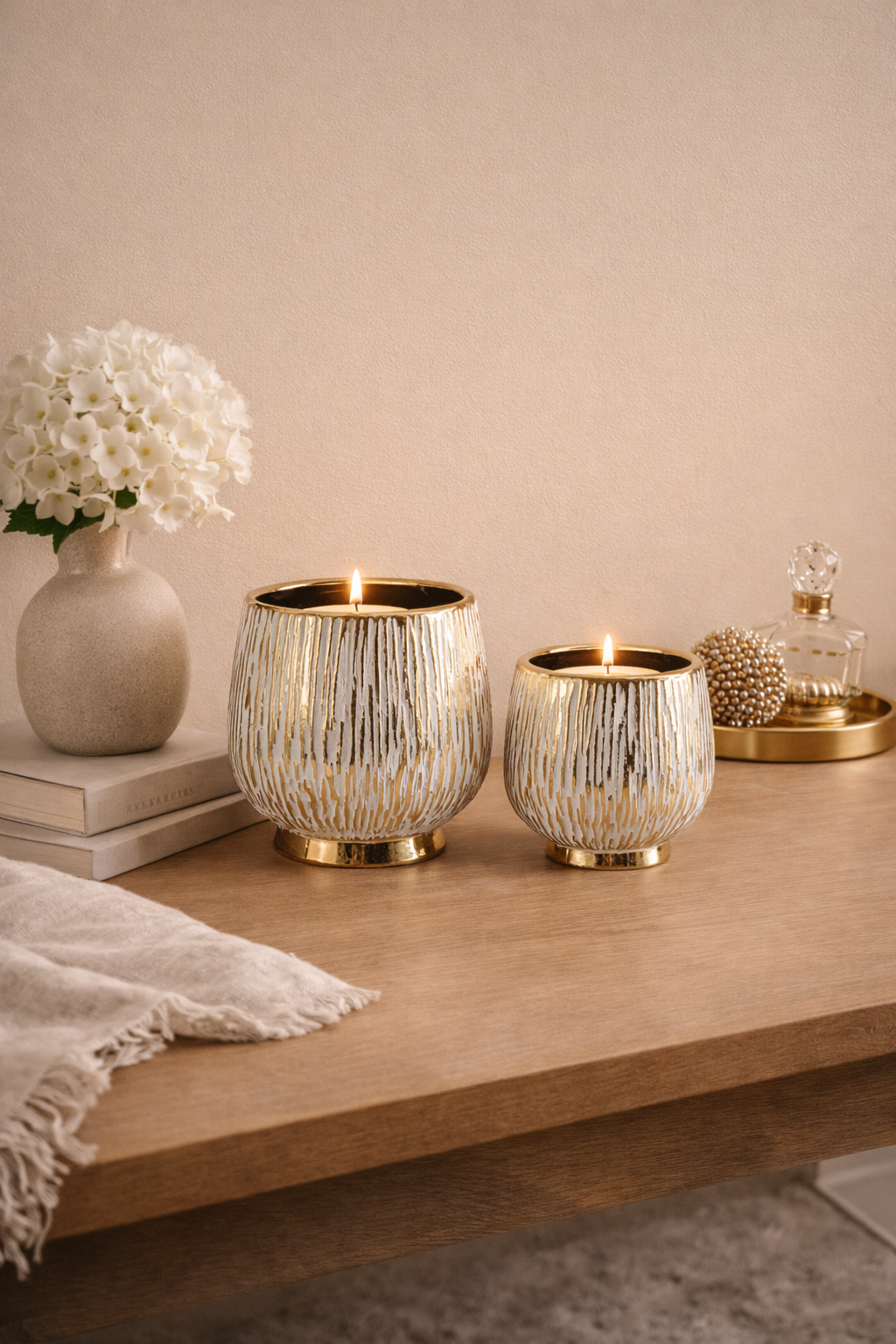 Two gold candle holders on a wooden surface with a vase of white flowers and other decorative items.