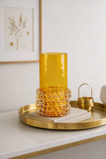 Yellow glass vase on a gold tray with a white wall and framed picture in the background