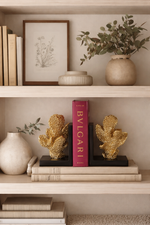 Decorative shelf with gold cactus sculptures, a pink Bulgari book, and a plant on a neutral background