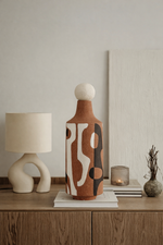 Decorative vase with abstract design on a wooden surface, with a lamp and candle in the background.