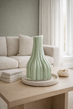 Green vase on a wooden table in a living room with a beige sofa and white curtains.