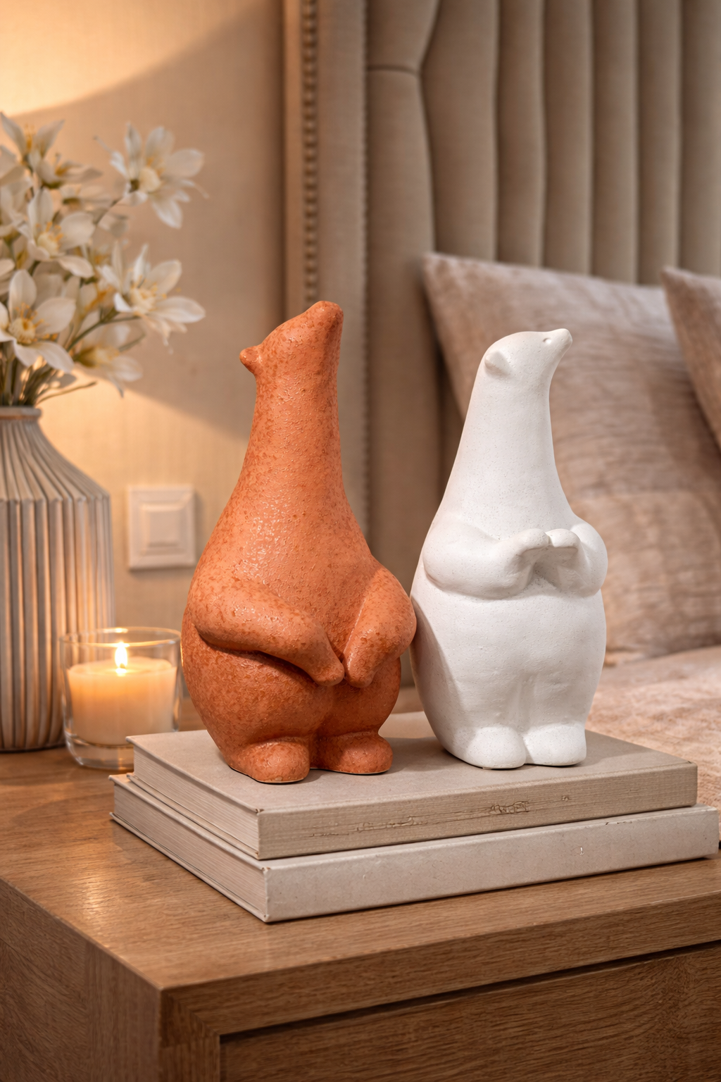 Two ceramic animal figurines, one orange and one white, on a wooden surface with a candle and flowers in the background.