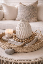 Decorative setting with a textured vase, candle, and books on a woven tray in a living room.