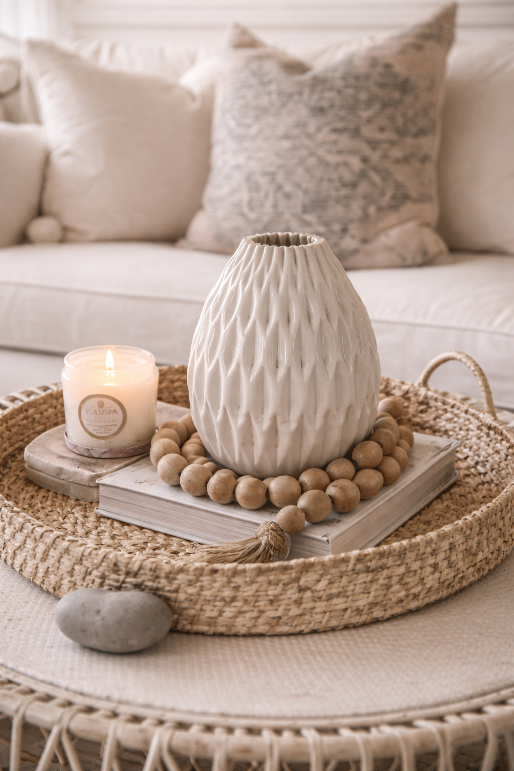 Decorative setting with a textured vase, candle, and books on a woven tray in a living room.