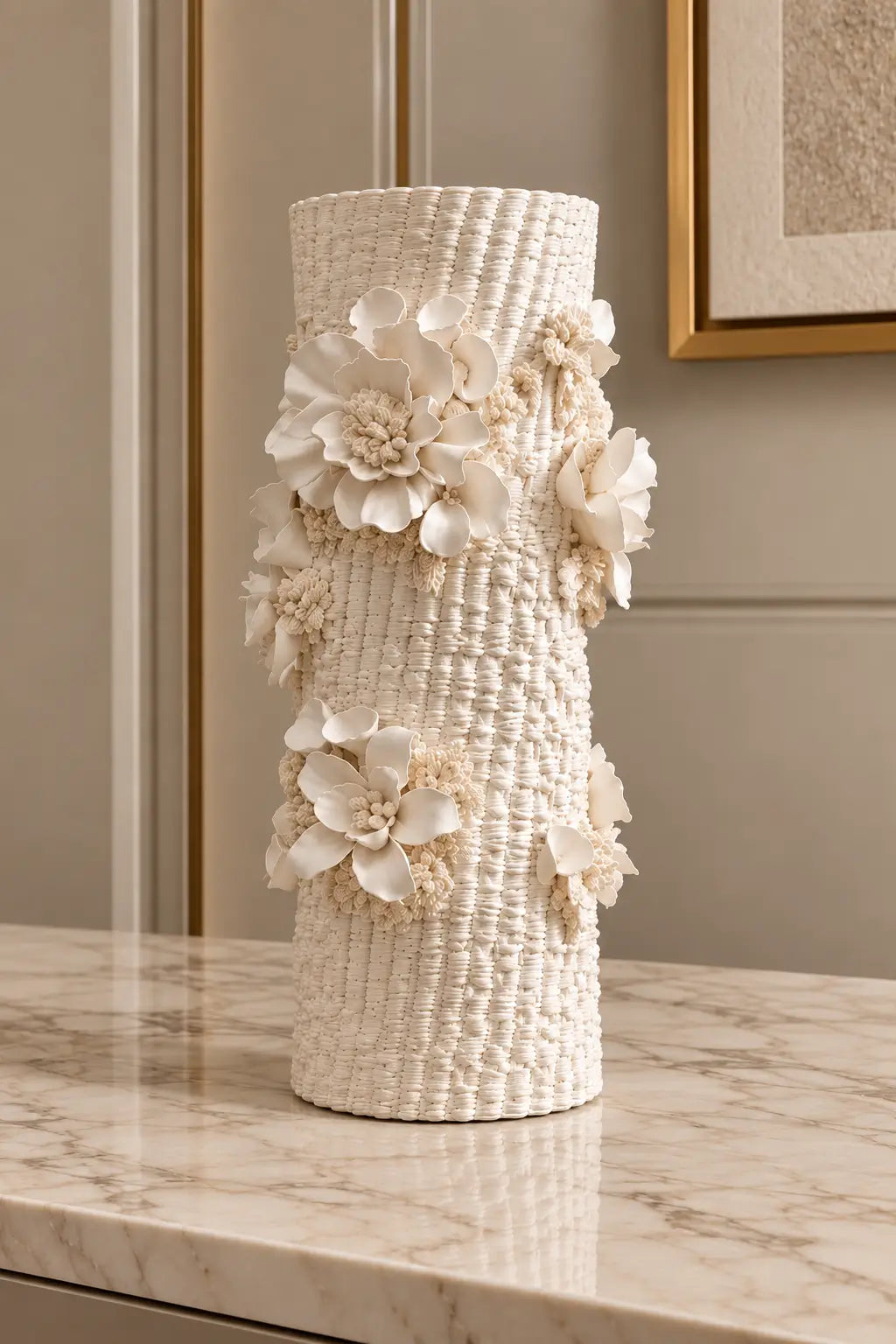 A tall cylindrical woven vase in warm ivory with large sculptural floral decorations, displayed on a marble surface against a beige interior wall.