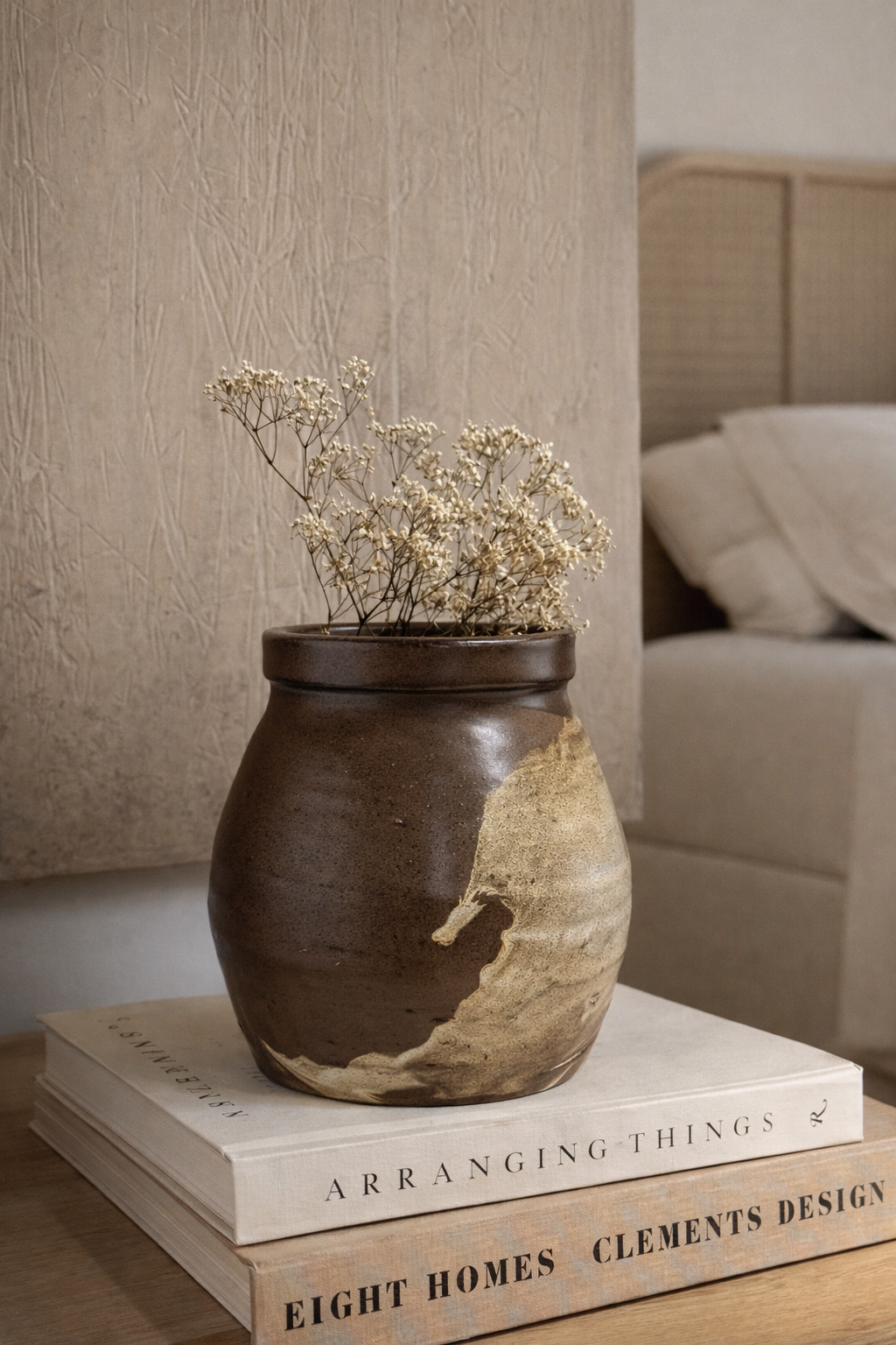 Brown ceramic vase with dried flowers on a stack of books in a room setting.