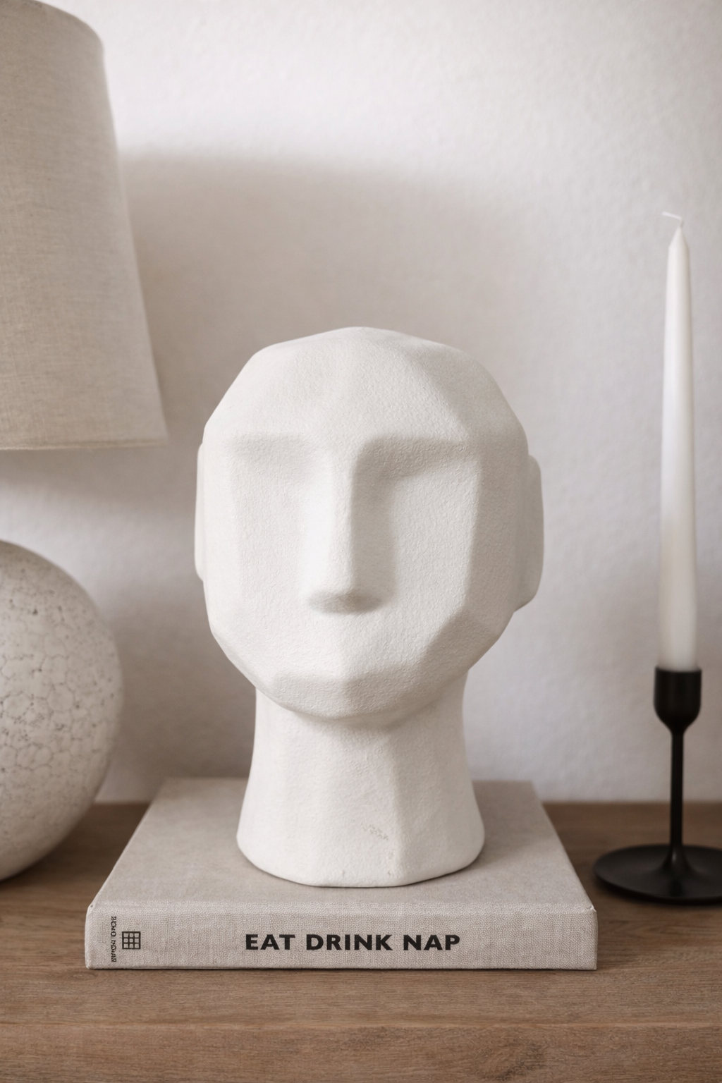 White marble head sculpture on a book with 'EAT DRINK NAP' text, next to a lamp and candle.