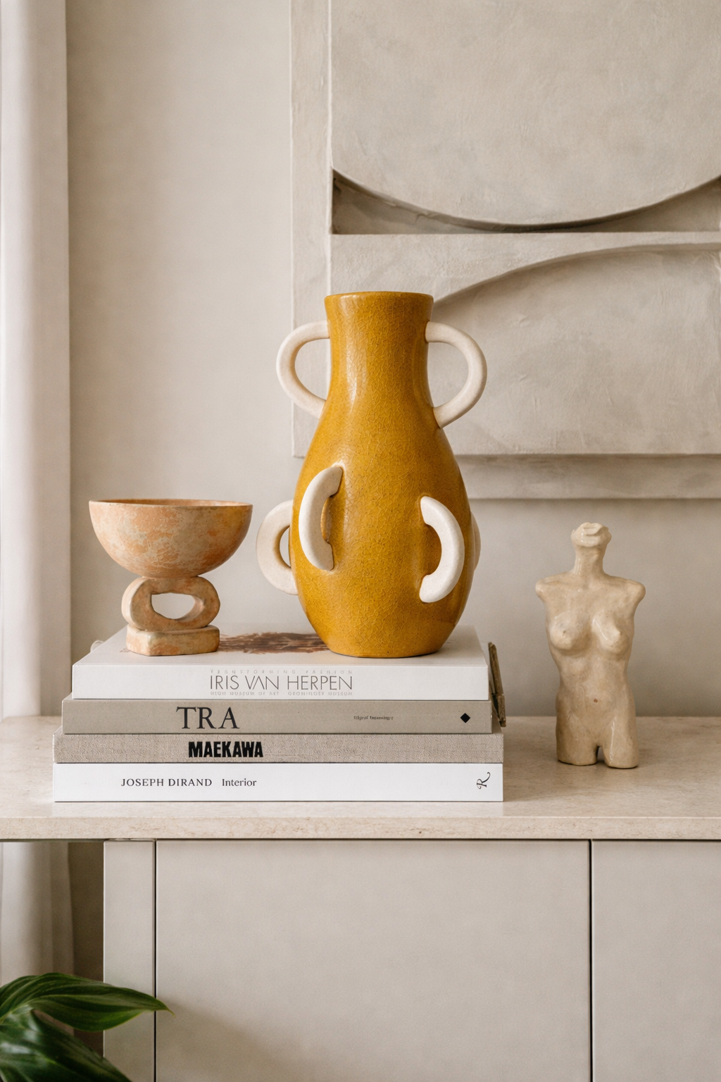 Yellow vase with white handles on books next to a small statue on a shelf.