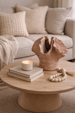 Round wooden coffee table with a ceramic vase, books, and a candle in a living room setting.