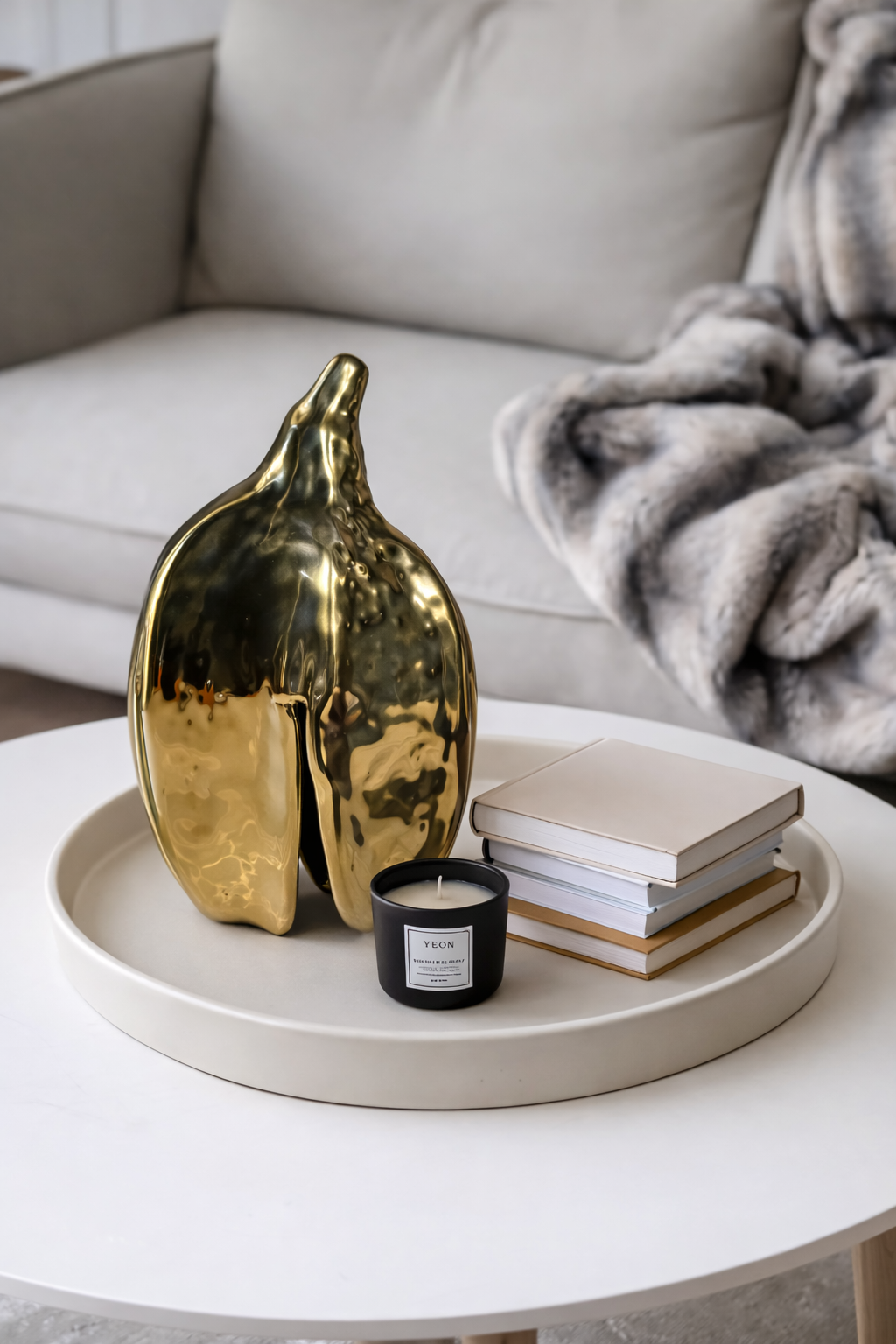 Decorative gold vase, black candle, and books on a white tray with a gray sofa in the background.