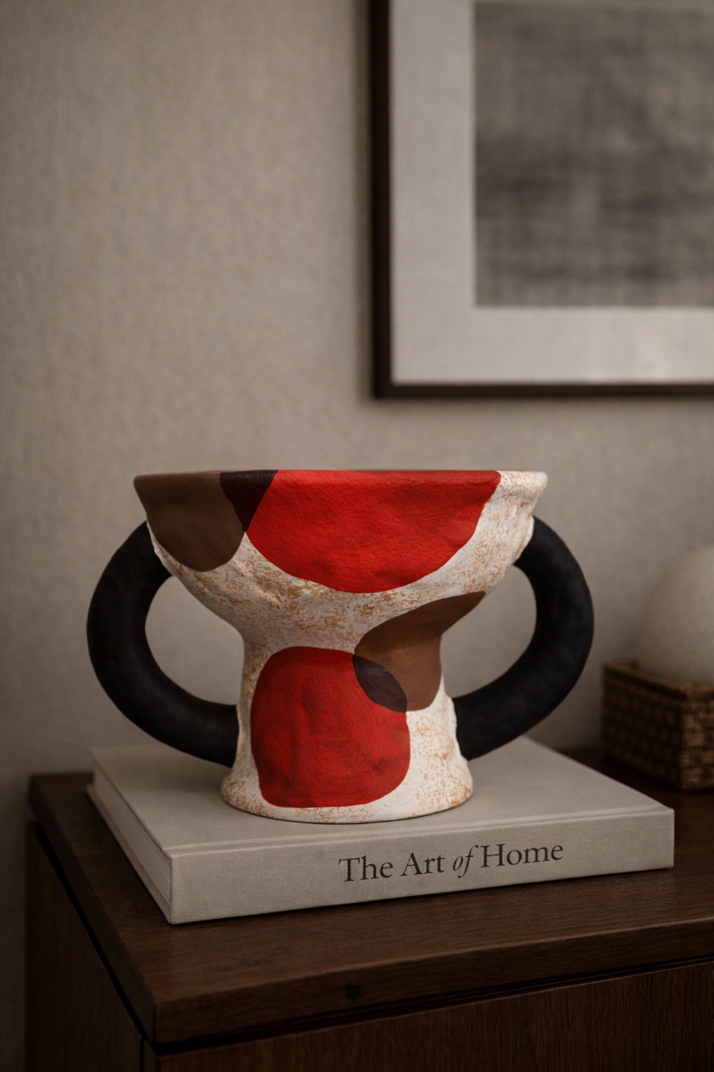 Ceramic vase with red, black, and beige design on a wooden surface with 'The Art of Home' book.